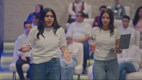 Two females are standing in the middle of an audience. One of them is holding a laptop and a drink