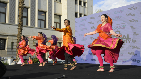 Dancers from Indian Embassy perform in colorful orange and pink saris on stage, arms spread