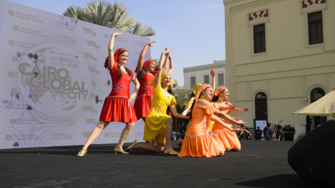 Female dancers from AUC Folklore smile mid-dance on stage with dresses in red, yellow, and orange.
