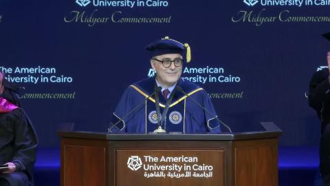 A man is wearing commencement cap and gown and speaking at a podium, he is wearing glasses. Text: The American University in Cairo. Midyear Commencement