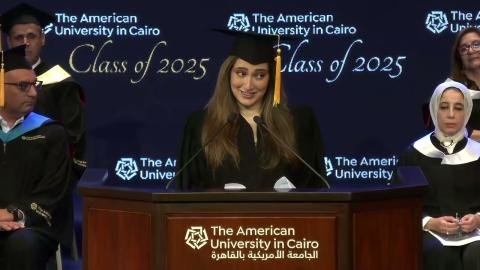 A woman is wearing a cap and gown. She is standing at a podium and talking into two microphone. There are males and females sitting behind her. Text: The American University in Cairo