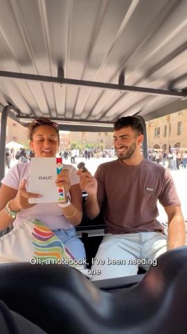 A male and female are sitting. He is holding a microphone and she is holding a colorful notebook written on it "AUC"