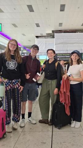 Four international students are standing and smiling. Some of them are holding luggage