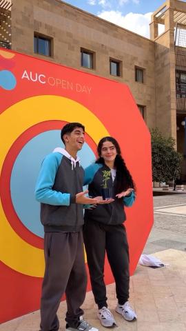 A male and a female standing in front of a colorful panel. Text: AUC Open Day
