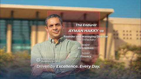 A man is standing and smiling. Text: Ayman Hakky &rsquo;08 &mdash; The Endurer.  Economics major. Founder and managing director of The TriFactory. AUC Driven by Excellence. Every Day.
