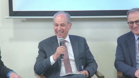 A man is smiling and talking into a microphone. He is wearing a suit. There is a man who is sitting next to him and also wearing a suit