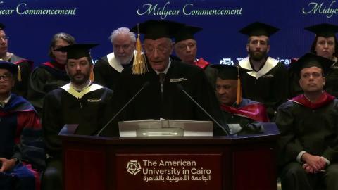 A man is standing at a podium, he is wearing a cap and gown. There are people sitting behind them, wearing caps and gowns