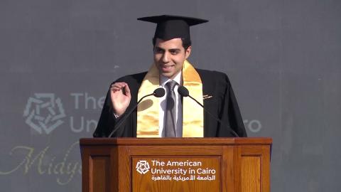 A male is talking at a podium and he is wearing a graduation cap and gown. Text: The American University in Cairo