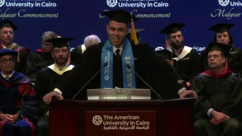 A man is standing at a podium, he is wearing a cap and gown. There are people sitting behind them, wearing caps and gowns