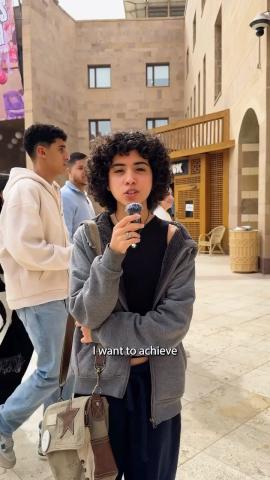 A female is standing and talking into a small microphone. There are people walking behind her