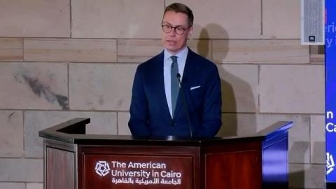 A male is standing a ta a podium and talking. Je is wearing a suit and glasses. Text: The American University in Cairo