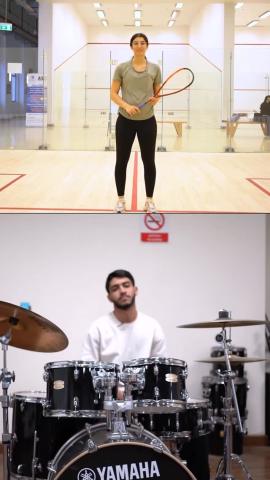 A female is standing, smiling and holding a squash racket inside a squash court. A male is sitting in front of a drum set