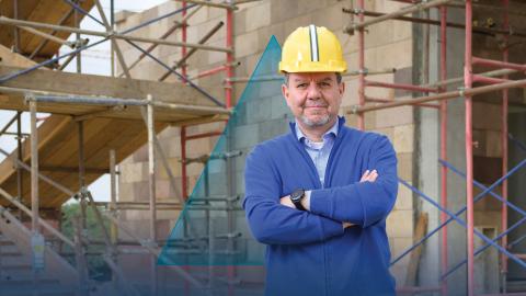A male is smiling and standing at a construction site. He is wearing a yellow helmet and blue overalls