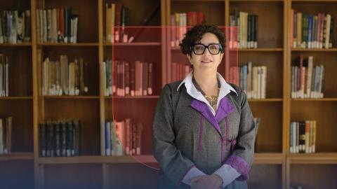 A female is standing and smiling. There is a bookshelf behind her.