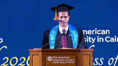 A male is wearing a cap and gown. He is standing at a podium and talking into a microphone. Text: The American University in Cairo