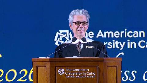 A man is wearing a cap and gown. He is standing at a podium and talking into a microphone. Text: The American University in Cairo