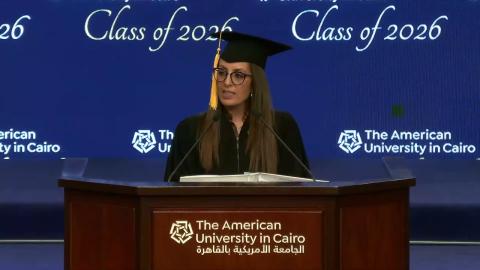 A woman is wearing a cap and gown. She is standing at a podium and talking into a microphone. Text: The American University in Cairo