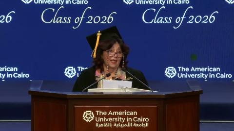 A woman is wearing a cap and gown. She is standing at a podium and talking into a microphone. Text: The American University in Cairo