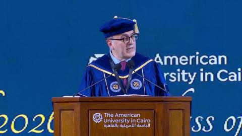 A man is wearing a cap and gown. He is standing at a podium and talking into a microphone. Text: The American University in Cairo
