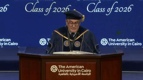 A man is wearing a cap and gown. He is standing at a podium and talking into a microphone. Text: The American University in Cairo