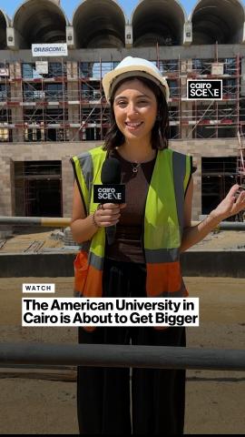 A female is wearing protective helmet and vest in construction site. She is standing and holding a microphone. Text: AUC is about to get bigger