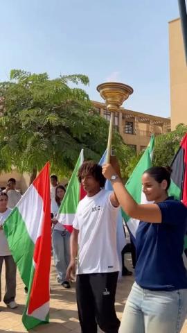 A male and female are holding a games torch. They are standing next to people holding flags