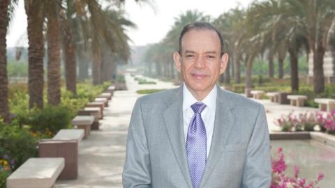 A man is standing in a garden and smiling. He is wearing a suit.