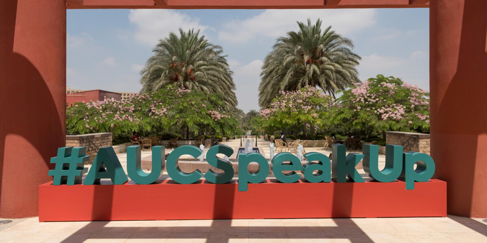A photo of a campus statue of the words "#AUCSpeakUp" in blue on a red rectangular box. There are trees and the AUC garden in the background and a red veranda/pillars on the sides and over the top.
