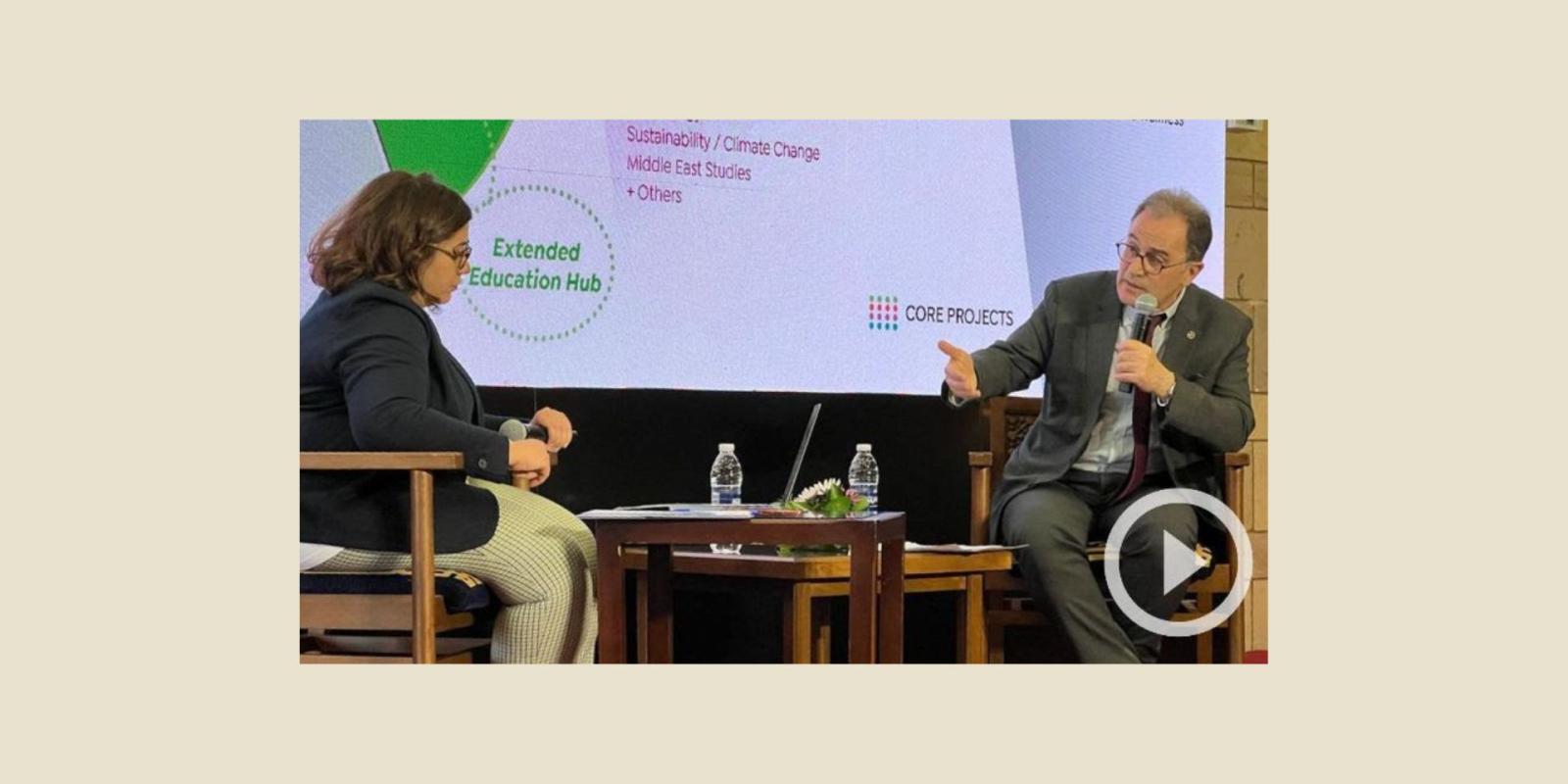 President Ahmad Dallal sits on a stage with moderator Amina Elbendary. He holds a microphone and gestures with his right hand, with a screen behind him showing an infographic explaining AUC's strategic priorities.