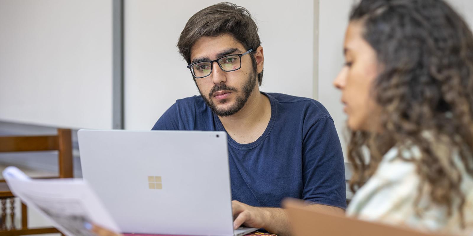 Boy in class with a laptop opened infront of him and a girl holding a paper