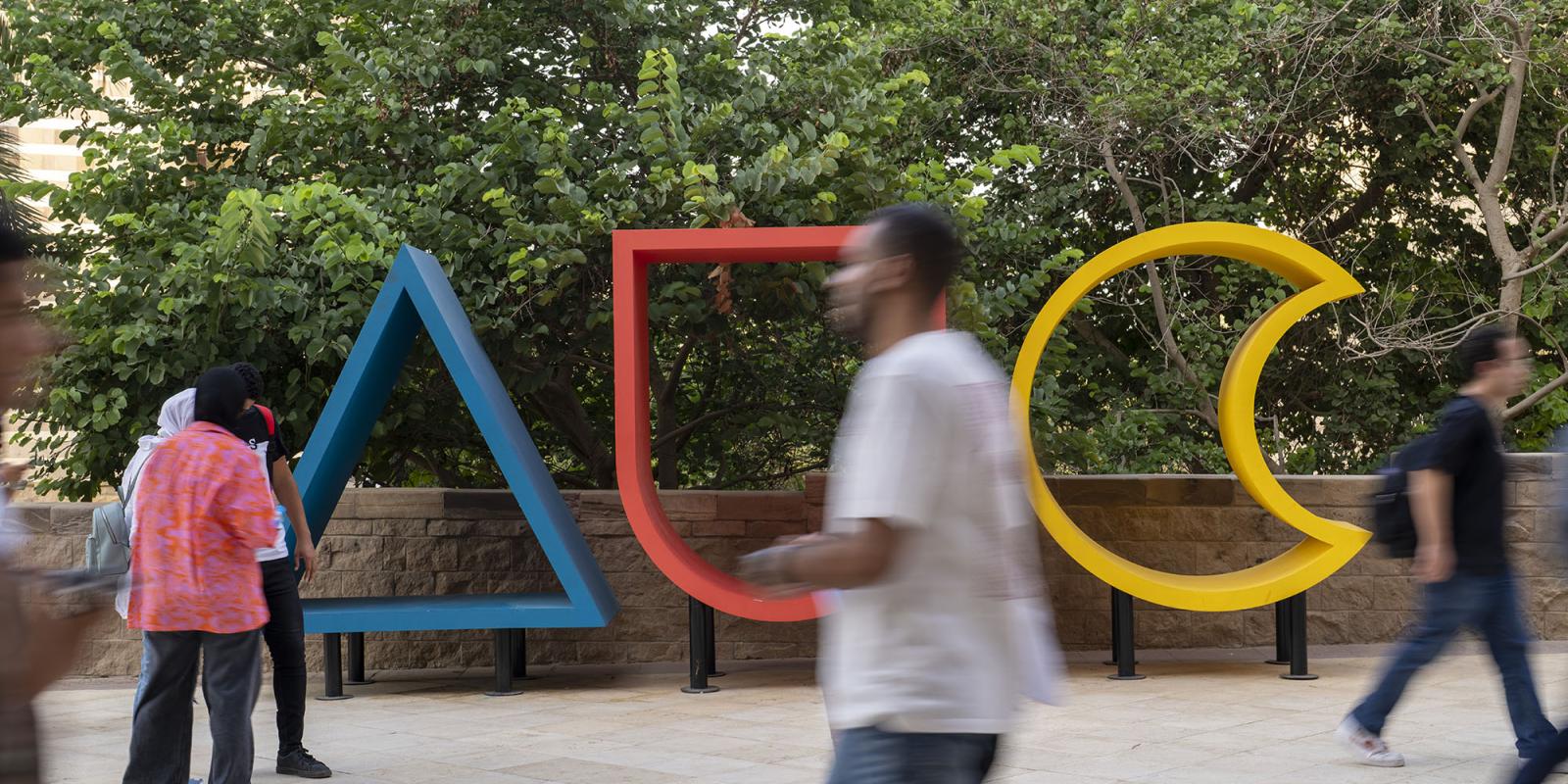 A large colorful wooden sign of AUC with students passing by