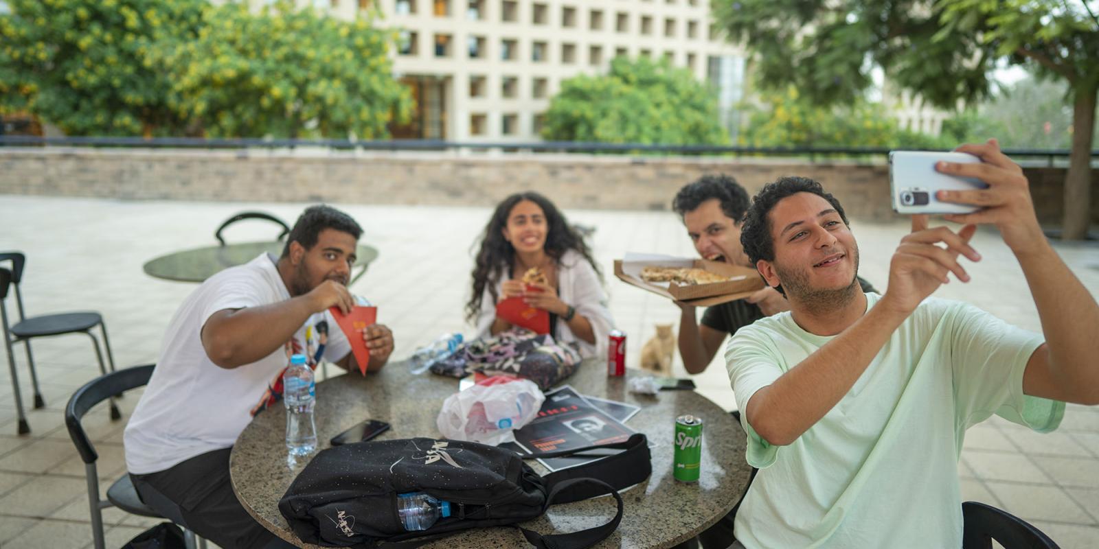Group of girls and boys sitting outdoors taking a selfie while eating