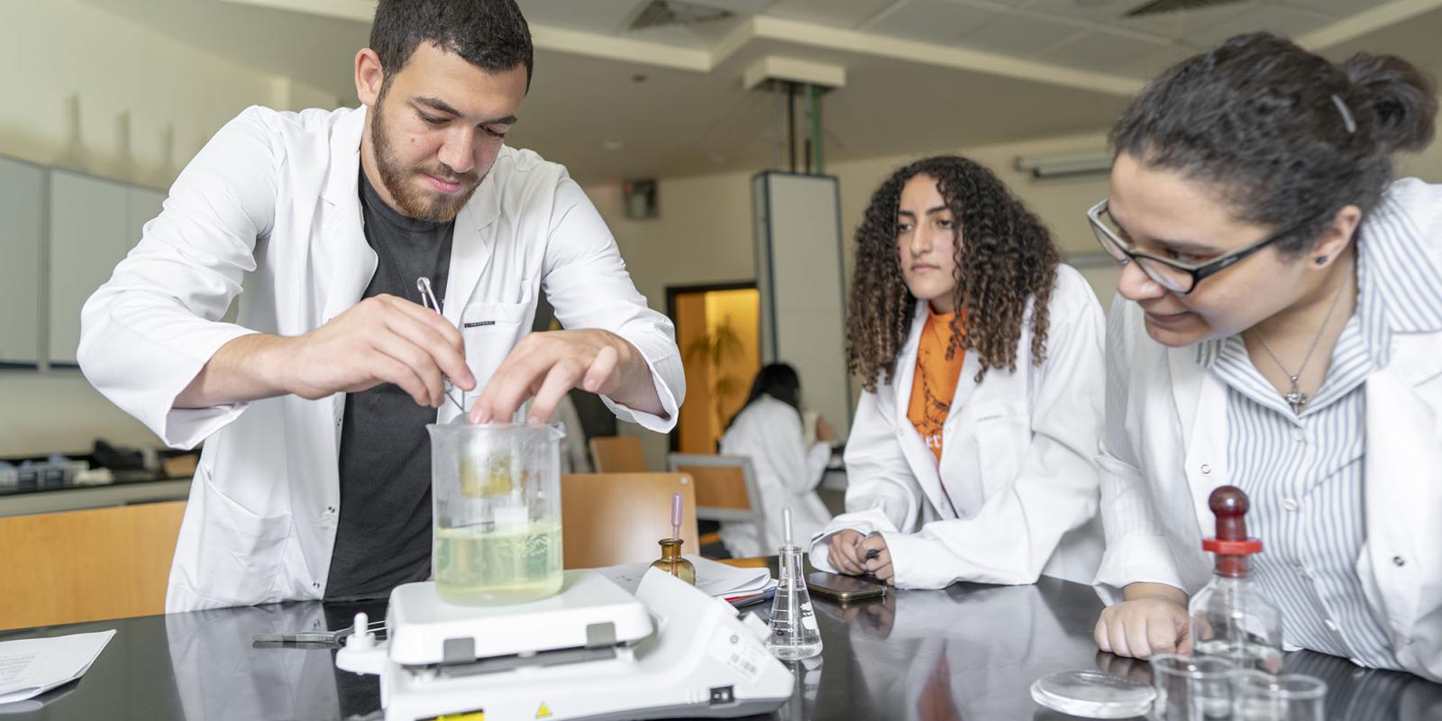Two girls and a boy in a lab wearing a white lab coat conducting an experiment with glass flasks and test tubes