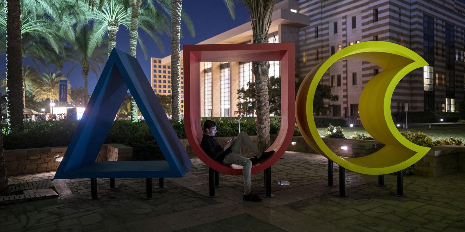 Big colored wooden AUC letters and student sitting in the letter U in front of a lit building at night