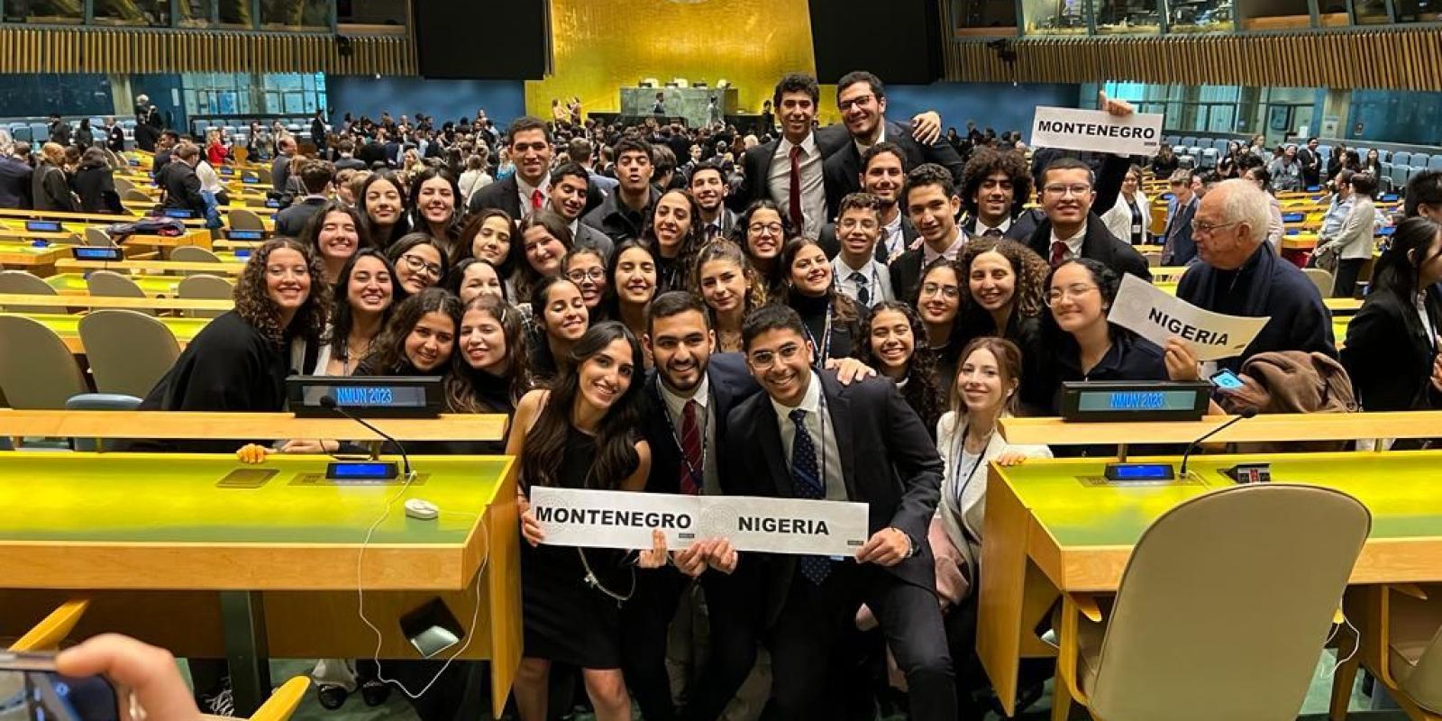 The CIMUN team stands wearing suits and professional dresses in an auditorium at the conference. Some students are holding signs saying "Montenegro" and "Nigeria"