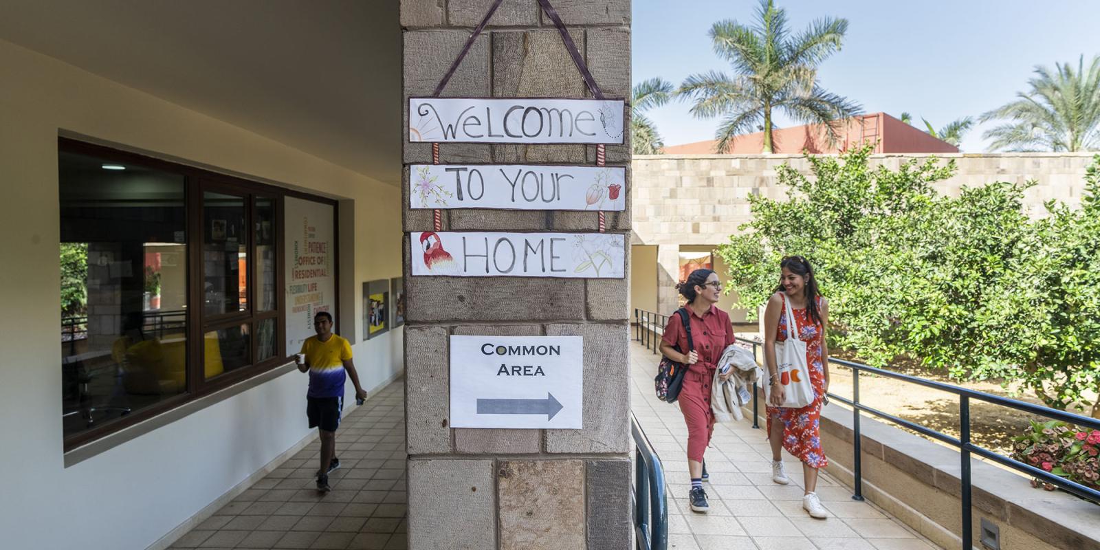 Two female students walking and talking together and a male student walking and holding a drinking cup, igns "Welcome to your home" and "Common area"