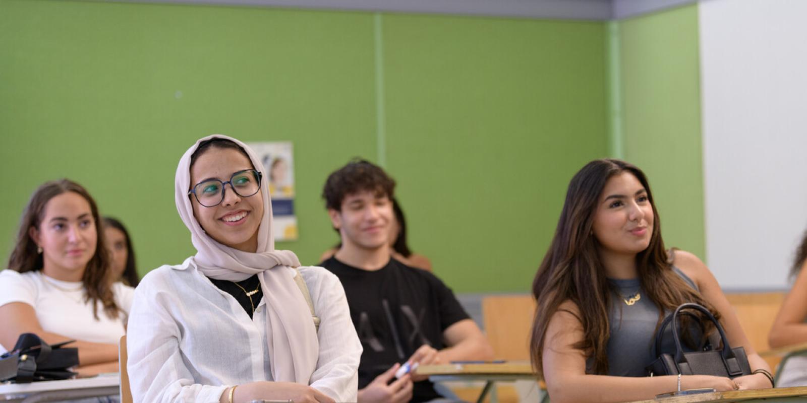 AUC students sit at desks in a classroom with a green wall behind them