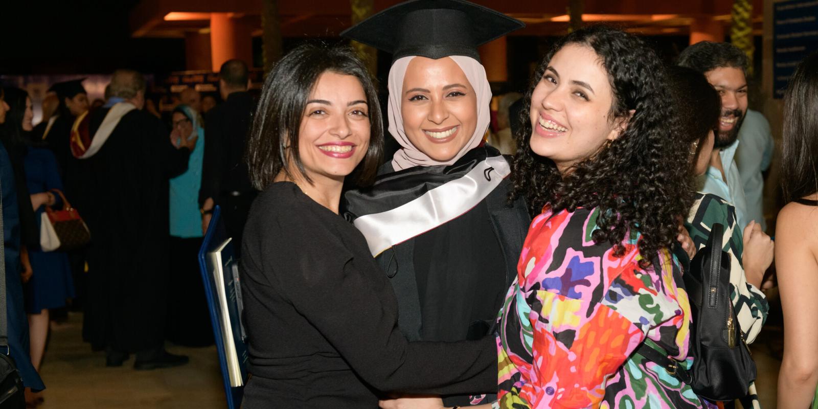 A graduating female student wearing a cap and gown with two other girls smiling
