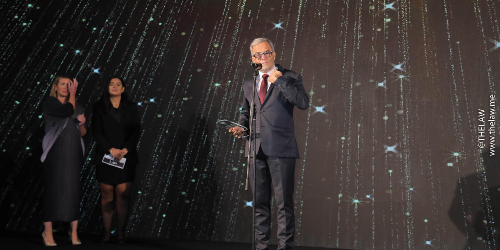 A man with a navy suit and a red tie stands in front of a conference background at a microphone, holding a glass award in his hand