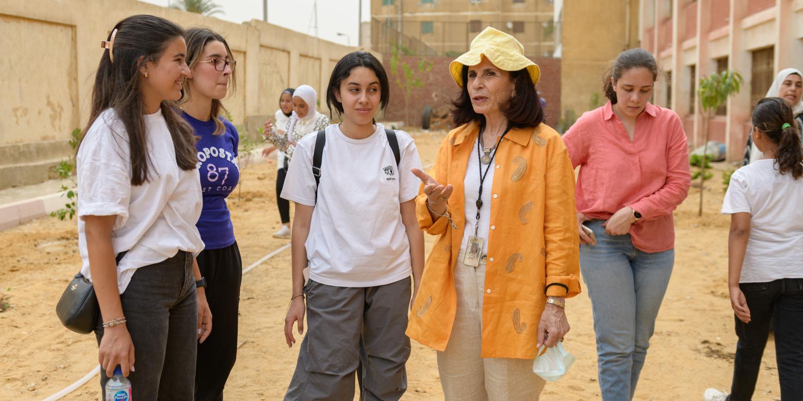 a woman in an orange shirt speaks with students in the yard of a school