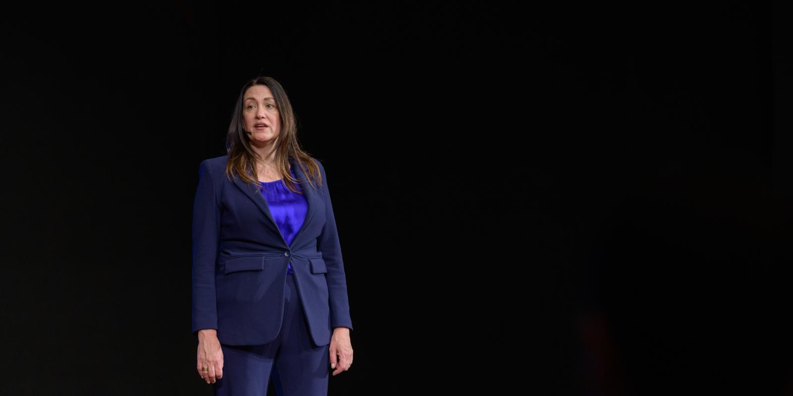 A woman presents in front of a dark background wearing a suit