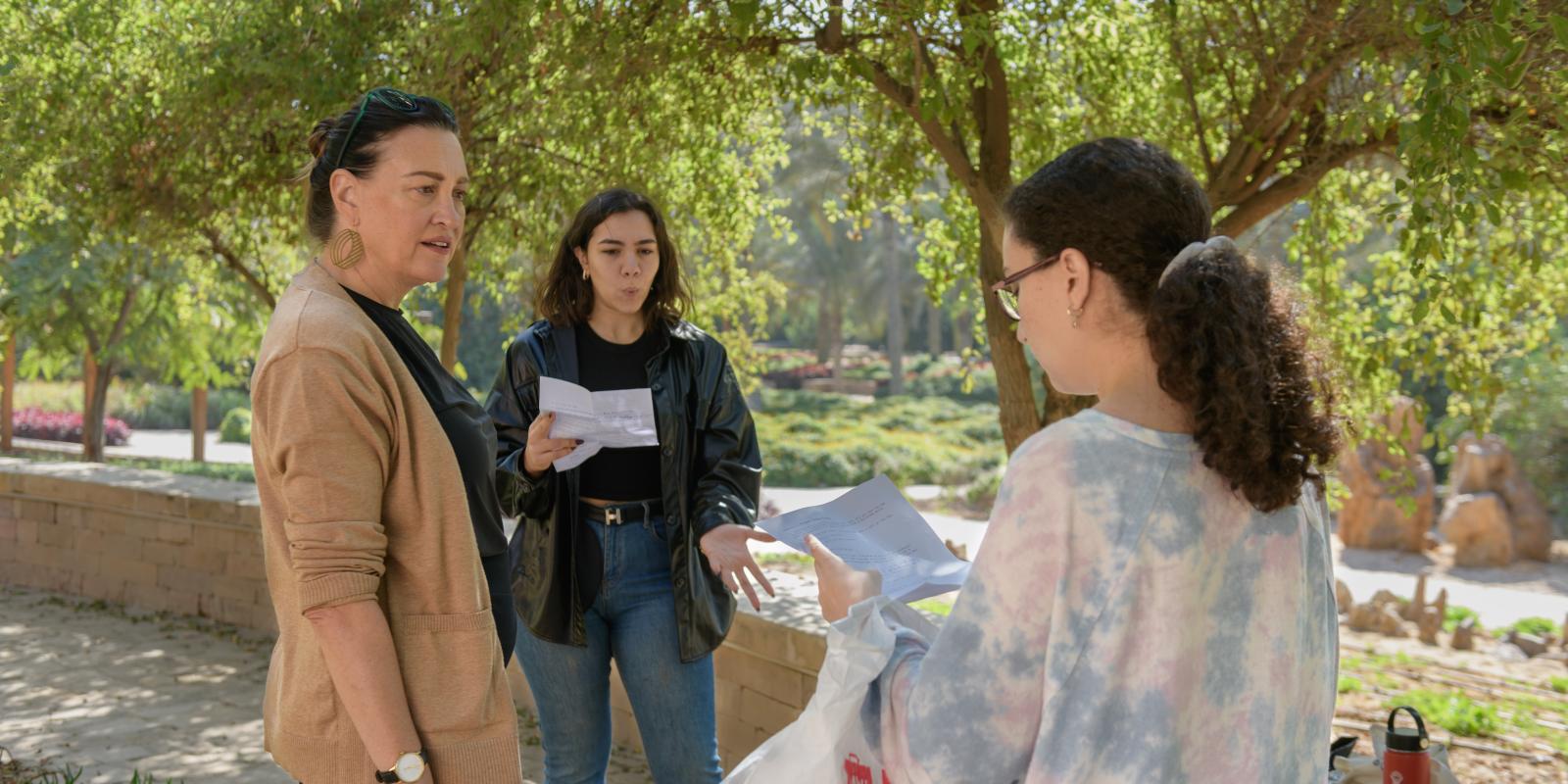A woman in a brown jacket works with students with scripts outdoors