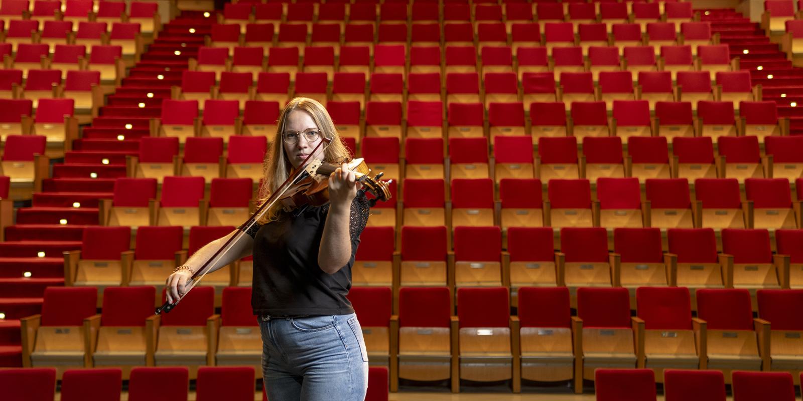 A female wearing glasses playing the violin in front of stage seats
