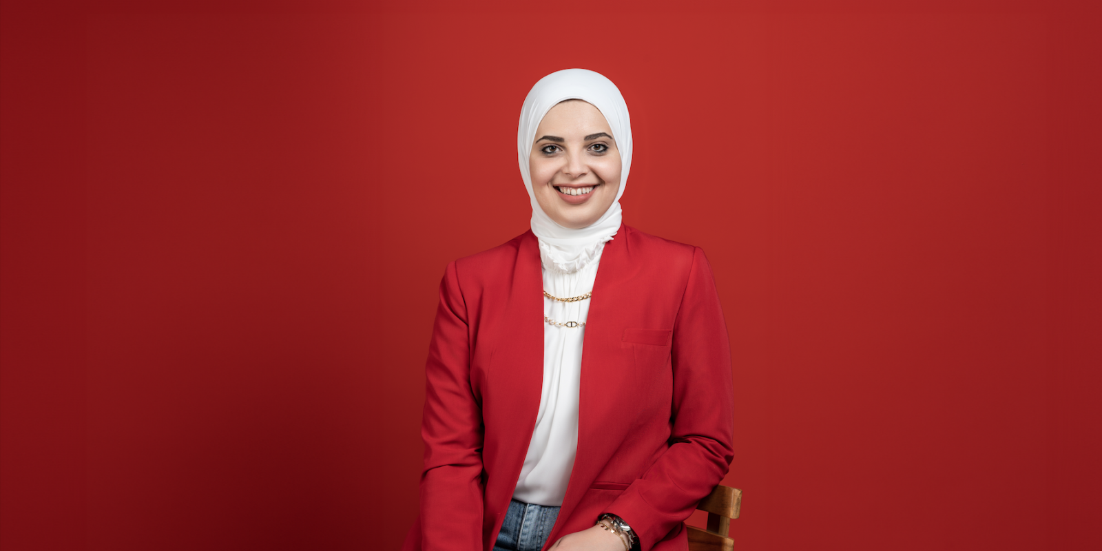 Woman in a red suit jacket and white hijab smiles in front of a red background