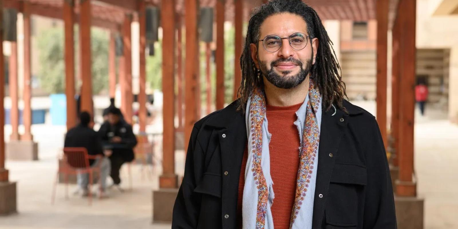 A bearded man is smiling and wearing glasses is standing outdoors