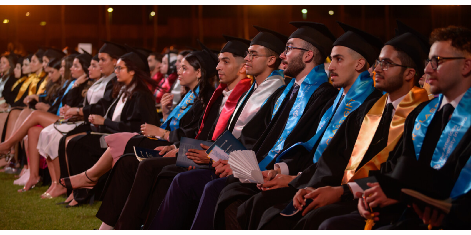 Students in caps and gowns sit in a row of chairs
