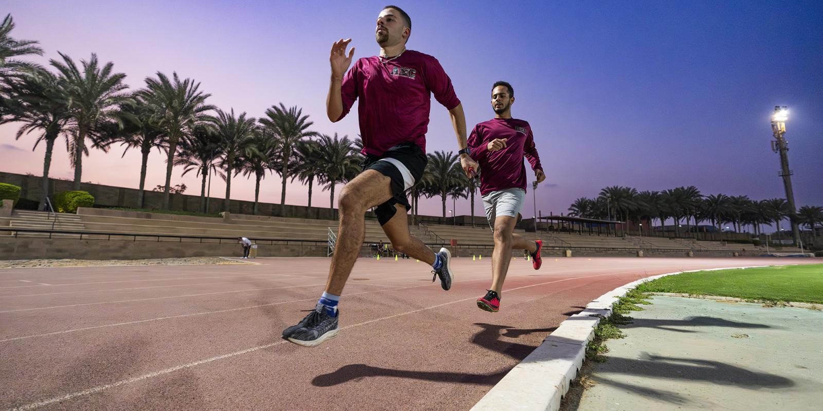 two male athletes running the track wearing maroon tshirts