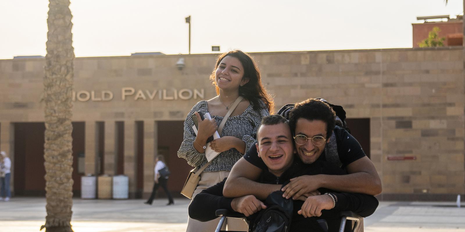 A boy sitting on a wheel chair smiling and behind him a boy and a girl standing