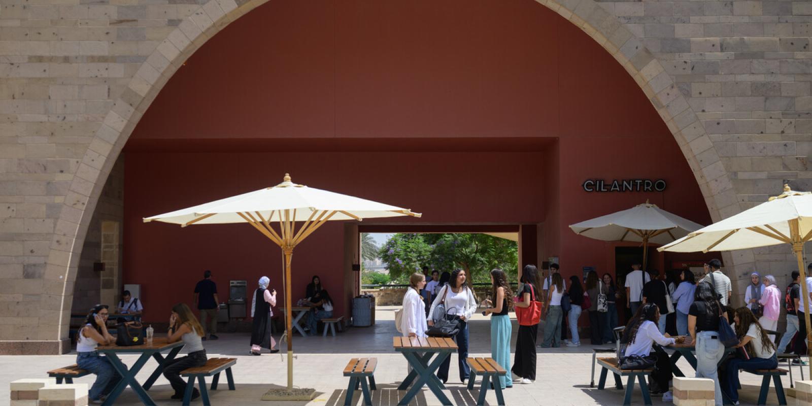 People sitting on benches and standing outdoors under shades talking and eating. There is a queue in front of a food outlet. Text: Cilantro