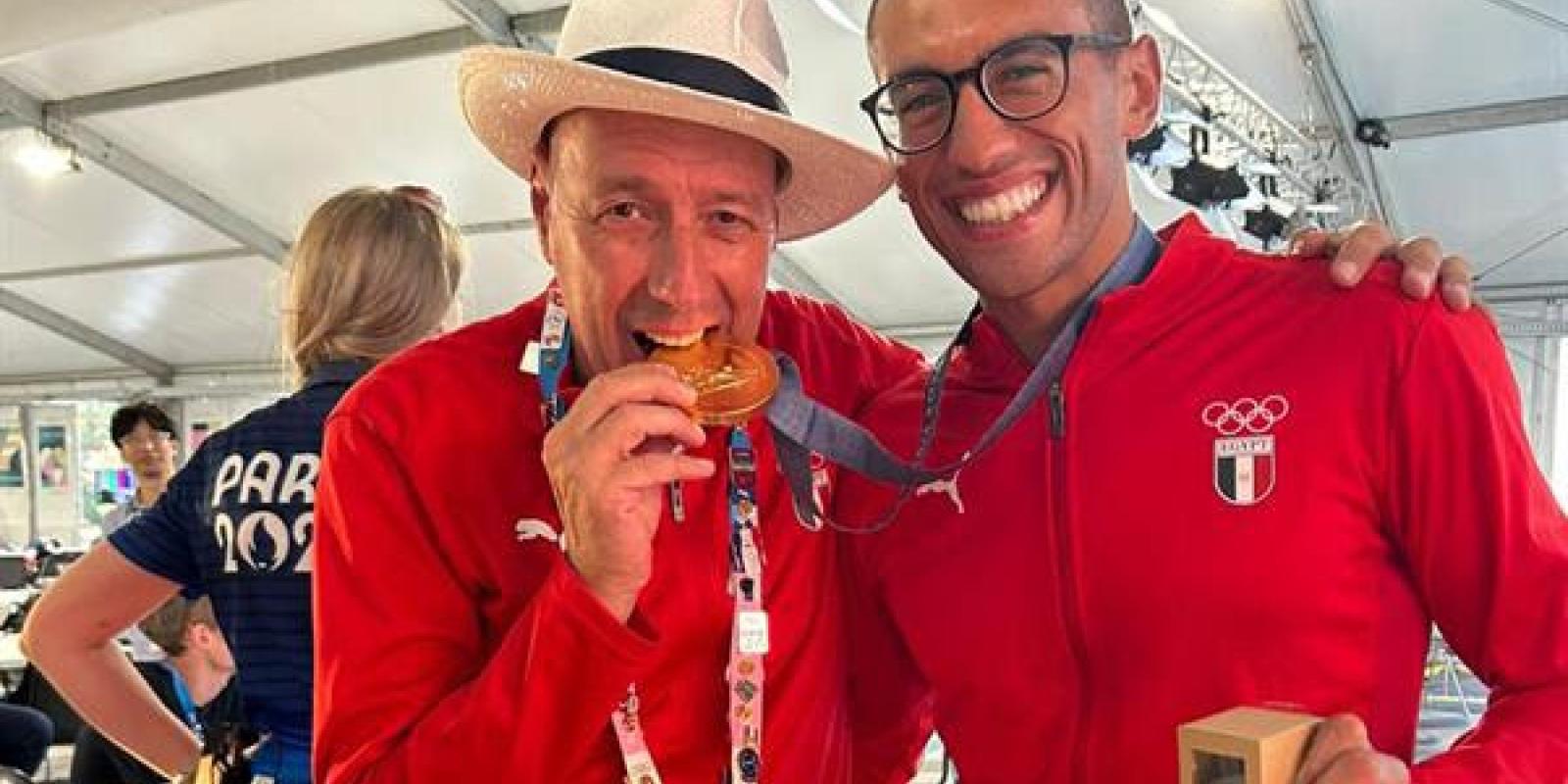 Two males are standing together, one is wearing a hat and holding a gold medal between his teeth, and the other is wearing glasses and holding a box